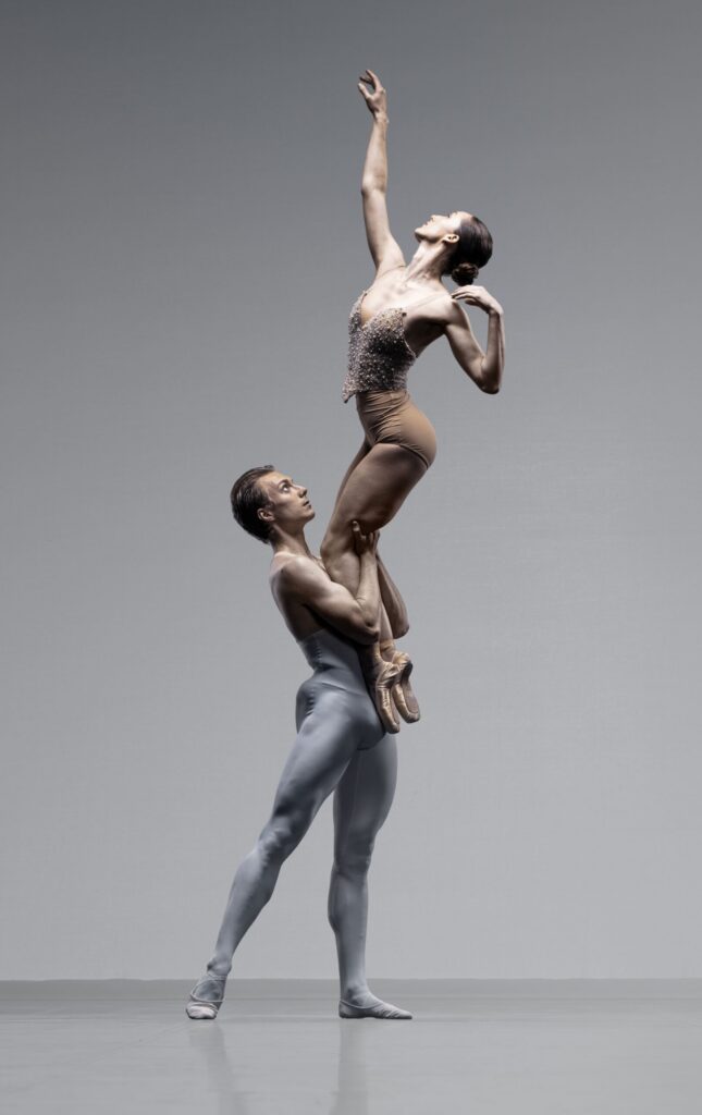 A male ballet dancer in gray lifts a female dancer in brown on pointe. She reaches upward with one arm, while he supports her with both hands. Both wear fitted costumes, posing gracefully against a plain gray background.
