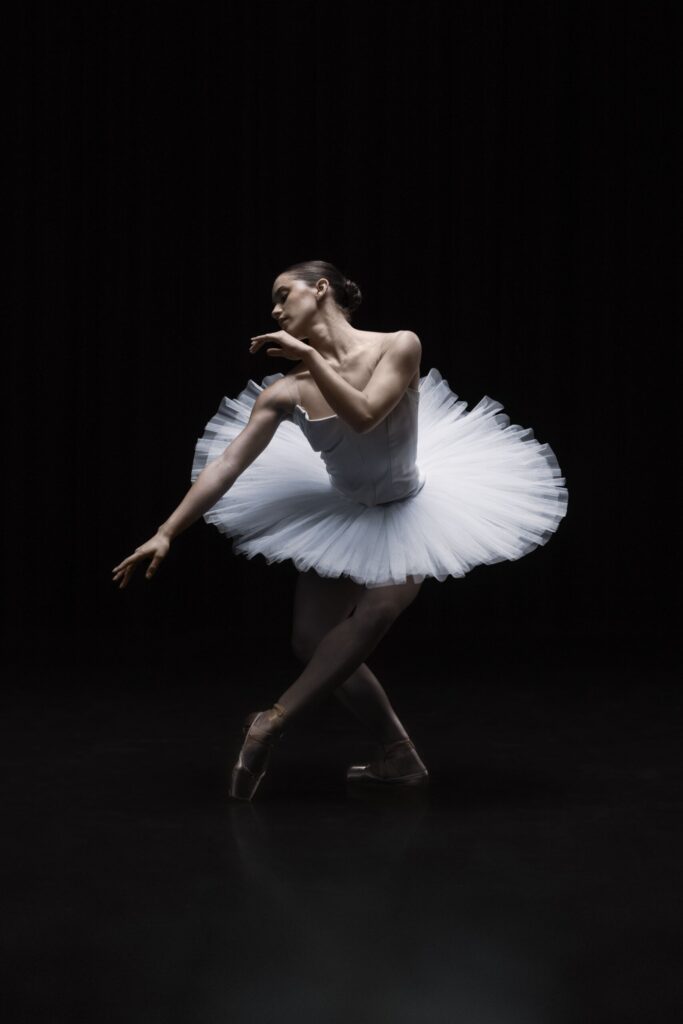 A ballerina in a white tutu dances gracefully on pointe against a dark background, her arms and body curved elegantly, with soft lighting highlighting her form.