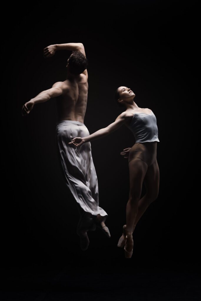 A male and female ballet dancer perform a leap together against a dark background, displaying strength and grace. The man wears white pants; the woman wears a white leotard and pointe shoes.