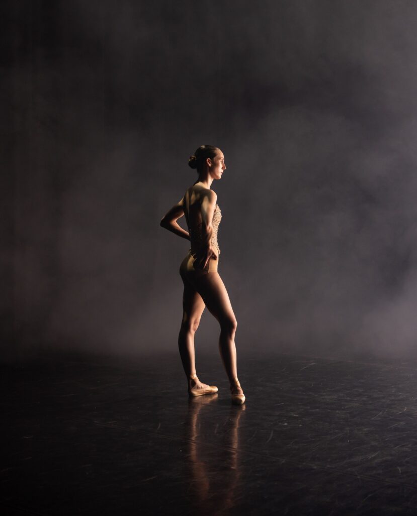 A ballerina stands on stage in a spotlight, wearing a leotard and ballet shoes, with her hands on her hips. The background is dark and smoky, highlighting her poised figure.