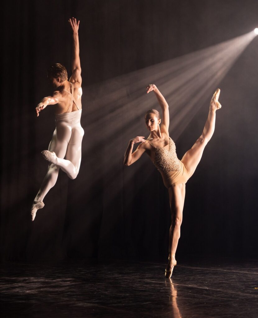 Two ballet dancers perform on stage: one leaps gracefully with an arm extended upward, while the other balances on one foot with the other leg lifted high. They are lit dramatically against a dark background.
