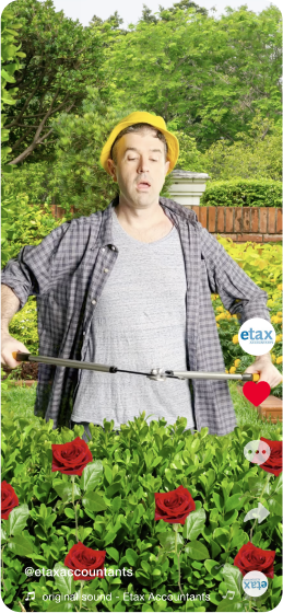 A man wearing a yellow hard hat and a plaid shirt, with a garden background. He is holding a garden hose and appears surprised. Red roses and green bushes are in the foreground. The image includes the Etax Accountants logo and a red heart icon on the right side.