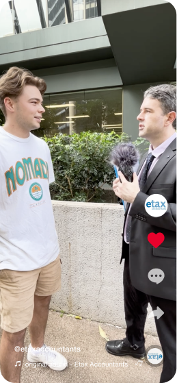 A young man wearing a white T-shirt with "NOMAD" printed on it being interviewed by a man in a suit holding a microphone. They are standing outside in front of a concrete wall with plants and a glass building in the background. Social media icons are visible on the side.