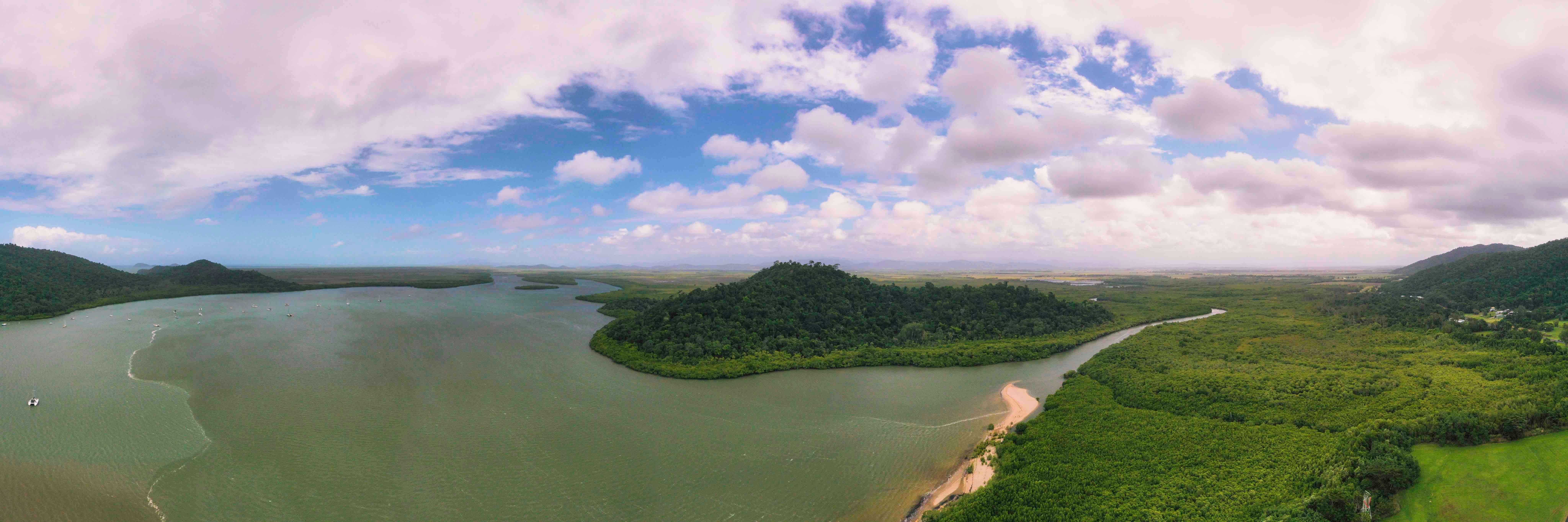 Aerial view of a coastal landscape featuring a winding river meeting the ocean, surrounded by lush greenery and forested hills. The sky is partly cloudy with patches of blue, creating a serene and natural atmosphere.