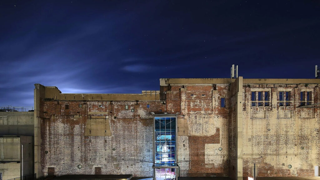 A large, weathered brick building with boarded windows, illuminated at night. A glass elevator shaft is centered, with light shining from inside. The sky is dark with faint, wispy clouds.
