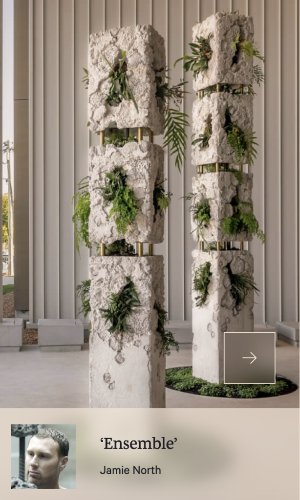 Two tall sculptural columns with rough, stone-like surfaces and embedded green plants stand indoors. The artwork is titled "Ensemble" by Jamie North.