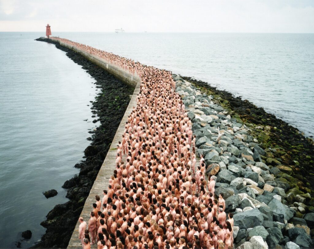 A large crowd of nude people stands closely together on a long stone pier stretching into the sea, with a red lighthouse at the far end under a cloudy sky.