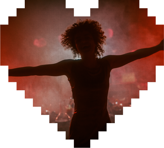 A person with curly hair stands with arms outstretched, silhouetted against red stage lights, framed by a pixelated heart shape.
