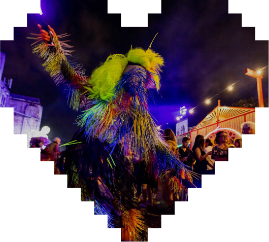 A person in a colorful, fringed costume dances at night under string lights, surrounded by people at an outdoor event. The image has a pixelated heart-shaped border.