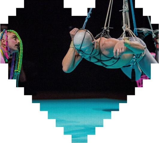 A person suspended in the air with rope harnesses performs an aerial act, while another person with colorful braided hair watches. The scene is framed by a pixelated heart shape.