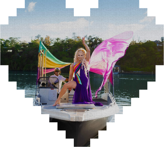 Person in a rainbow sash and flowing purple dress poses confidently on a boat with pride flags, while another person sits behind the wheel. The image is framed in a heart shape.
