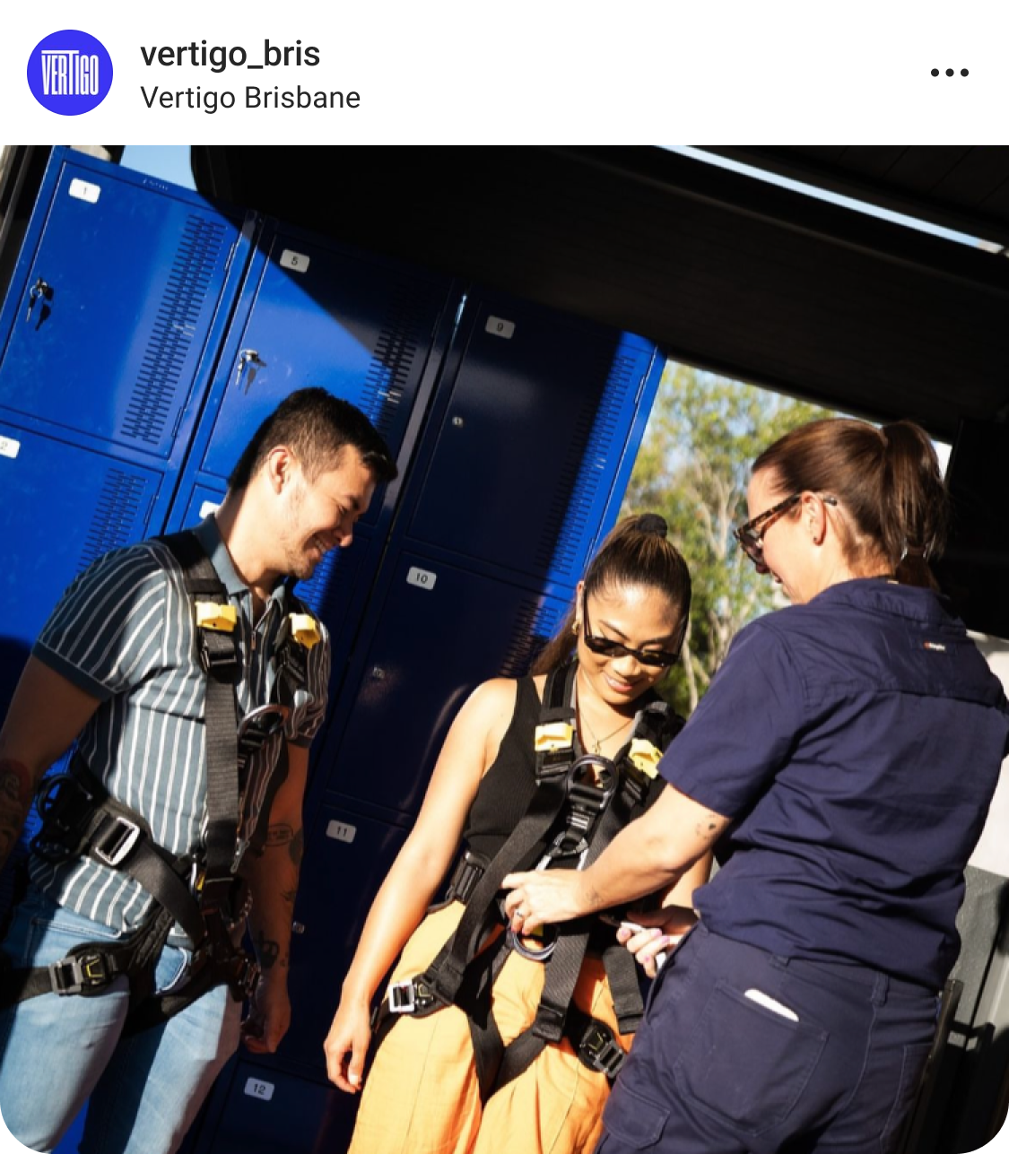 Two people wearing harnesses stand next to a staff member in uniform, who is adjusting the harness for the woman in sunglasses. Blue lockers are visible in the background.