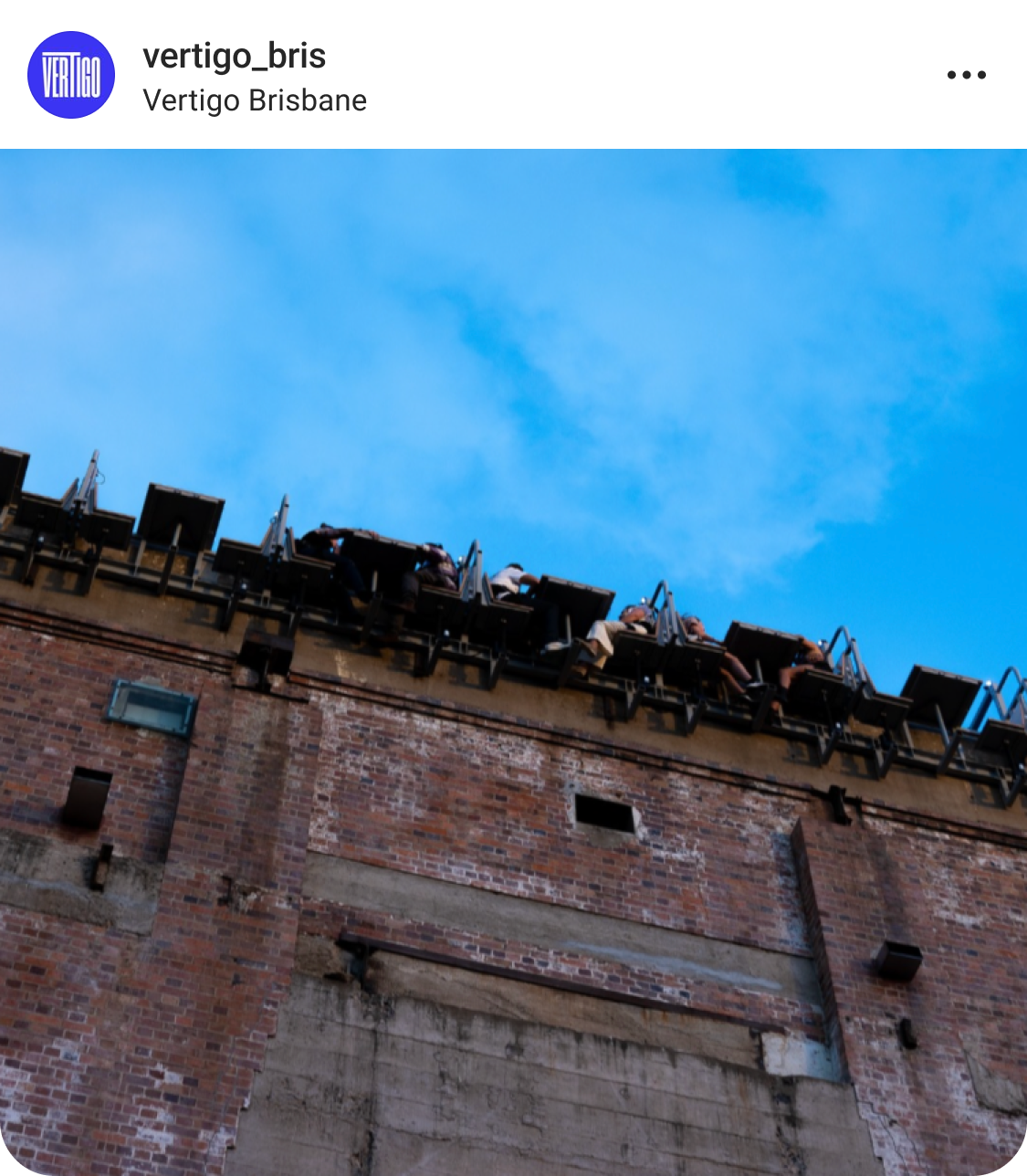 A view looking up at people seated on the edge of a tall brick structure against a blue sky, likely participating in an adventure or activity at Vertigo Brisbane.