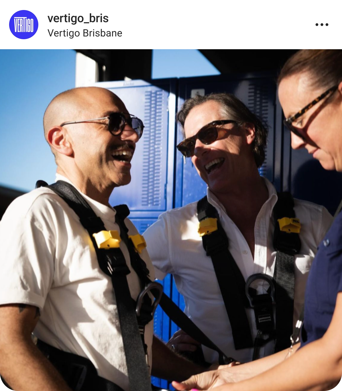 Two people in harnesses smile and laugh together while a third person helps adjust their gear. The scene appears outdoors in bright daylight.