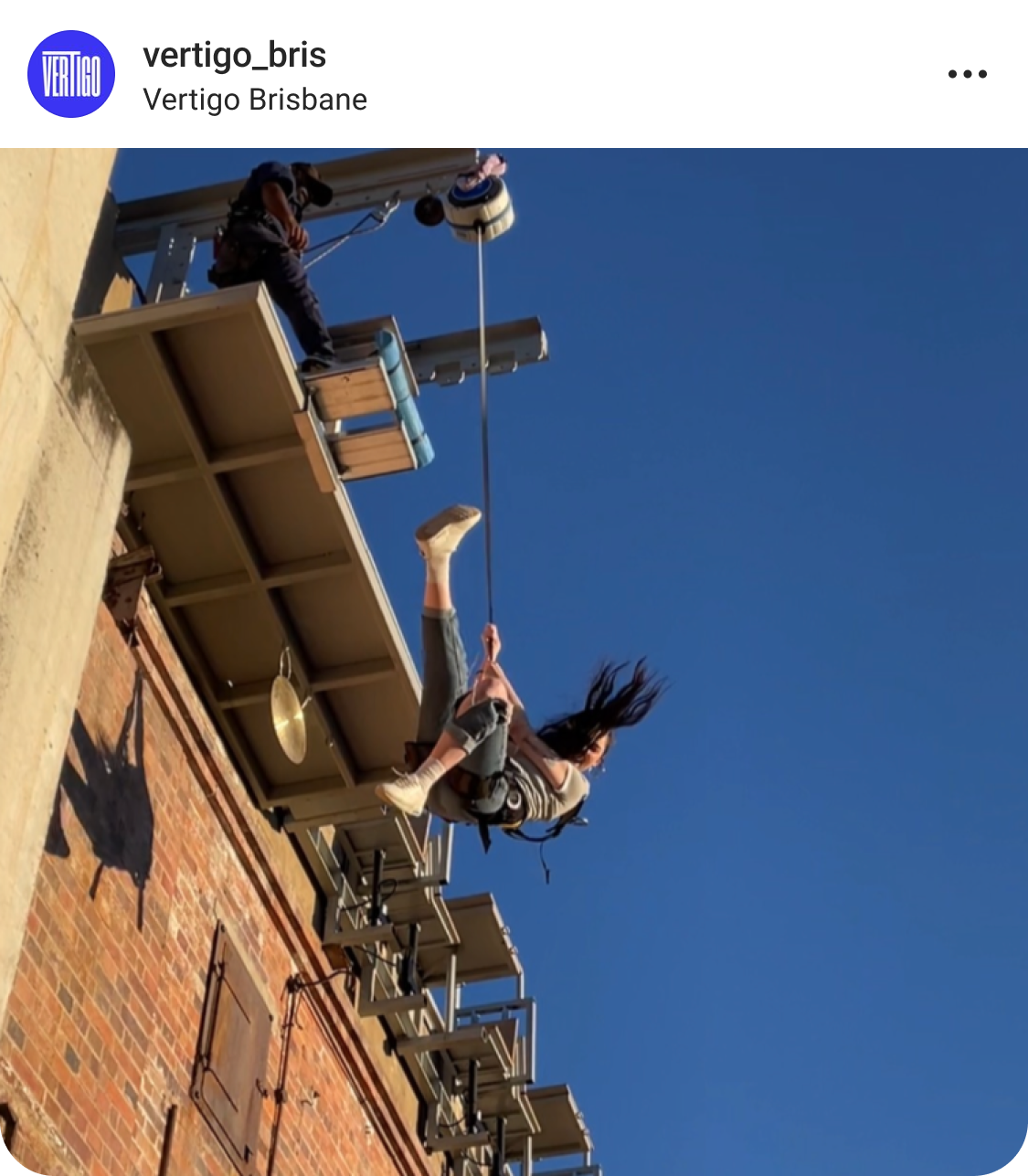 A person swings on a harness attached to a high platform against a clear blue sky, with a brick building and equipment visible in the background.