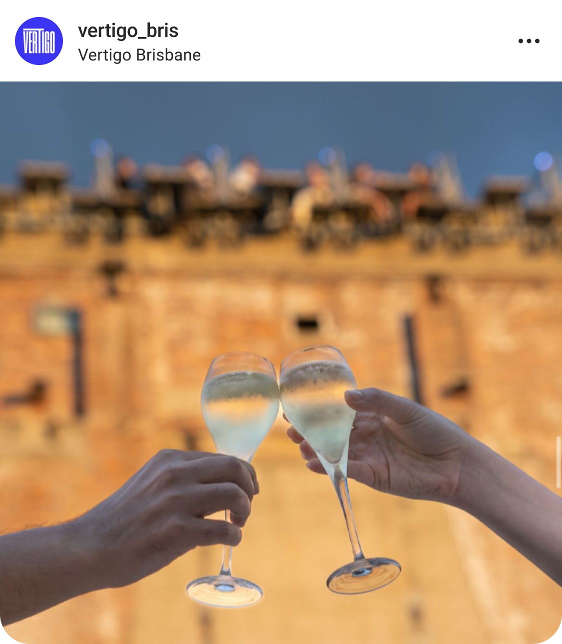 Two hands clink glasses of white wine or champagne in front of a blurred brick building, with people visible at the top of the structure against a blue sky.