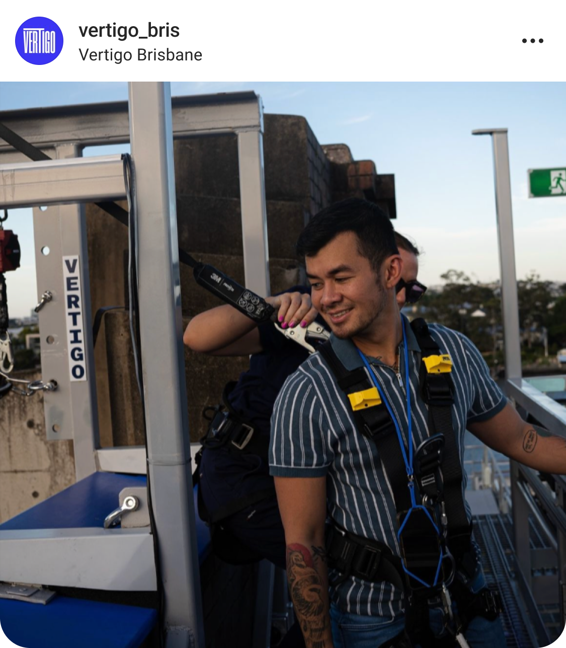 A man in a striped shirt and safety harness stands on an outdoor platform, smiling as another person checks his gear. A Vertigo Brisbane sign and equipment are visible in the background.