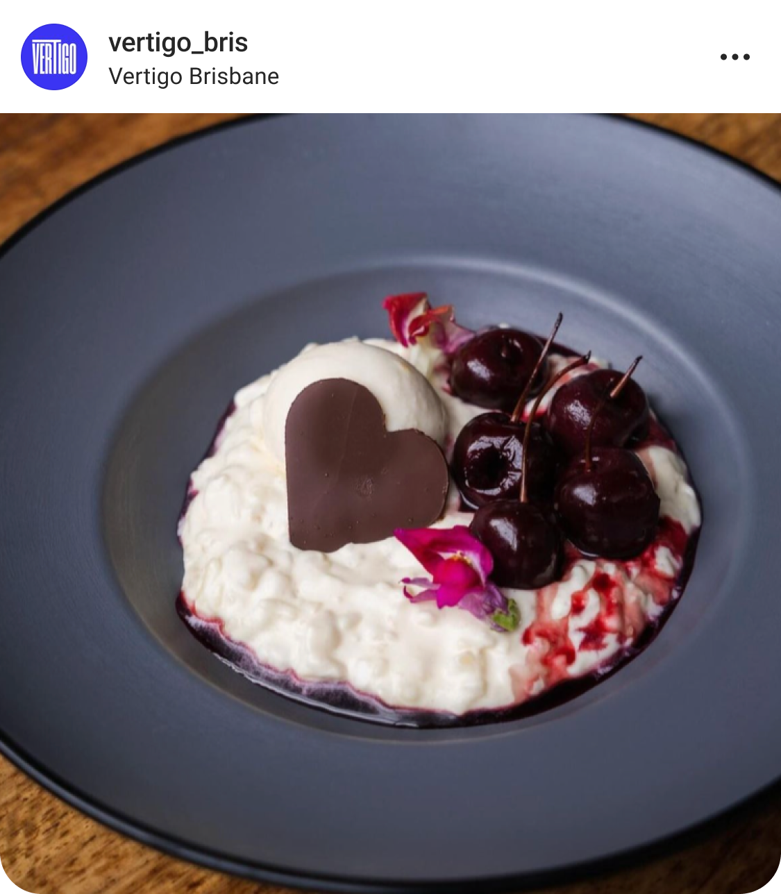 A black plate with creamy rice pudding, dark cherries, a scoop of ice cream, a chocolate heart, and edible pink flowers, served on a wooden table.