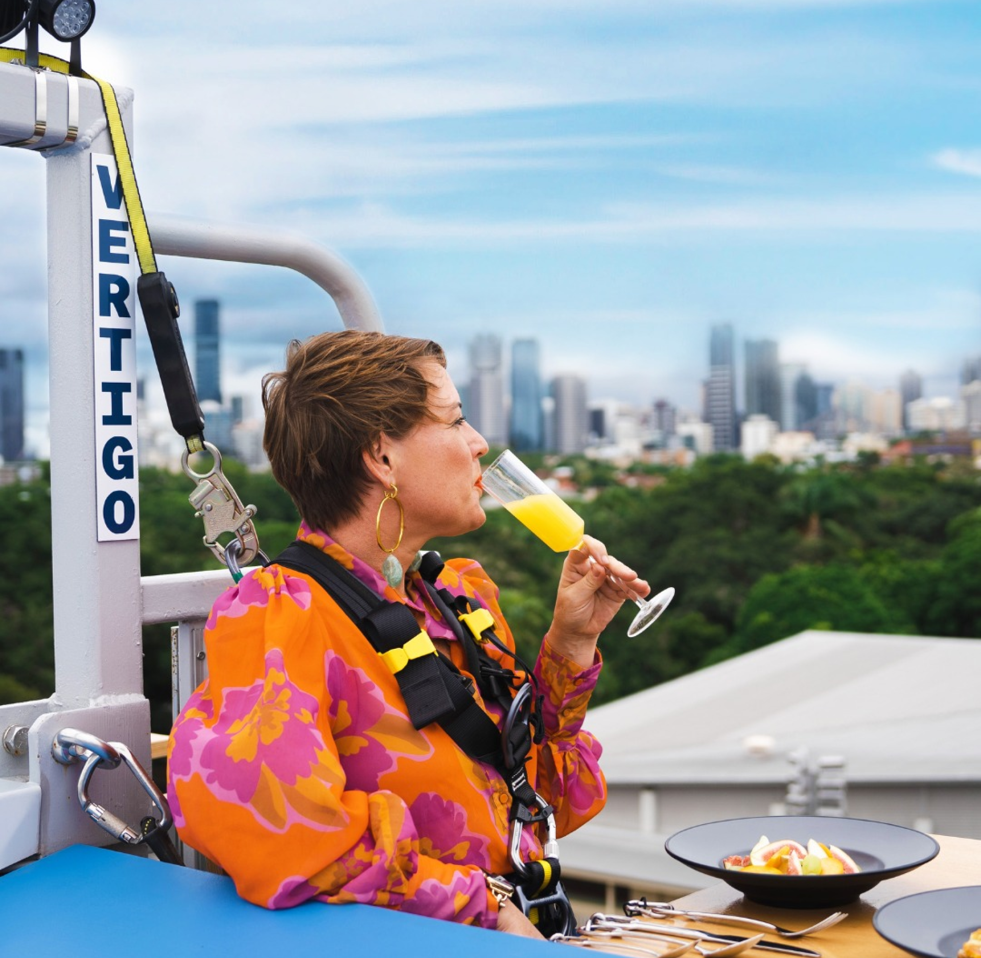 A person in a harness sips a mimosa at a rooftop dining setup, with a city skyline and green trees in the background. The word "VERTIGO" is visible on a safety rail beside them.