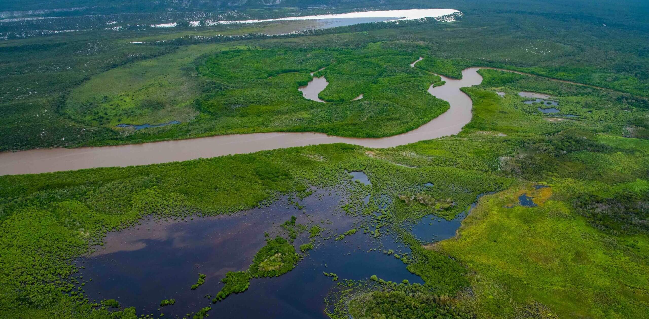 Aerial view of a winding river surrounded by lush green vegetation. There are multiple bodies of water, including a prominent blue lake in the foreground. The landscape is expansive, with distant hills under a clear sky.