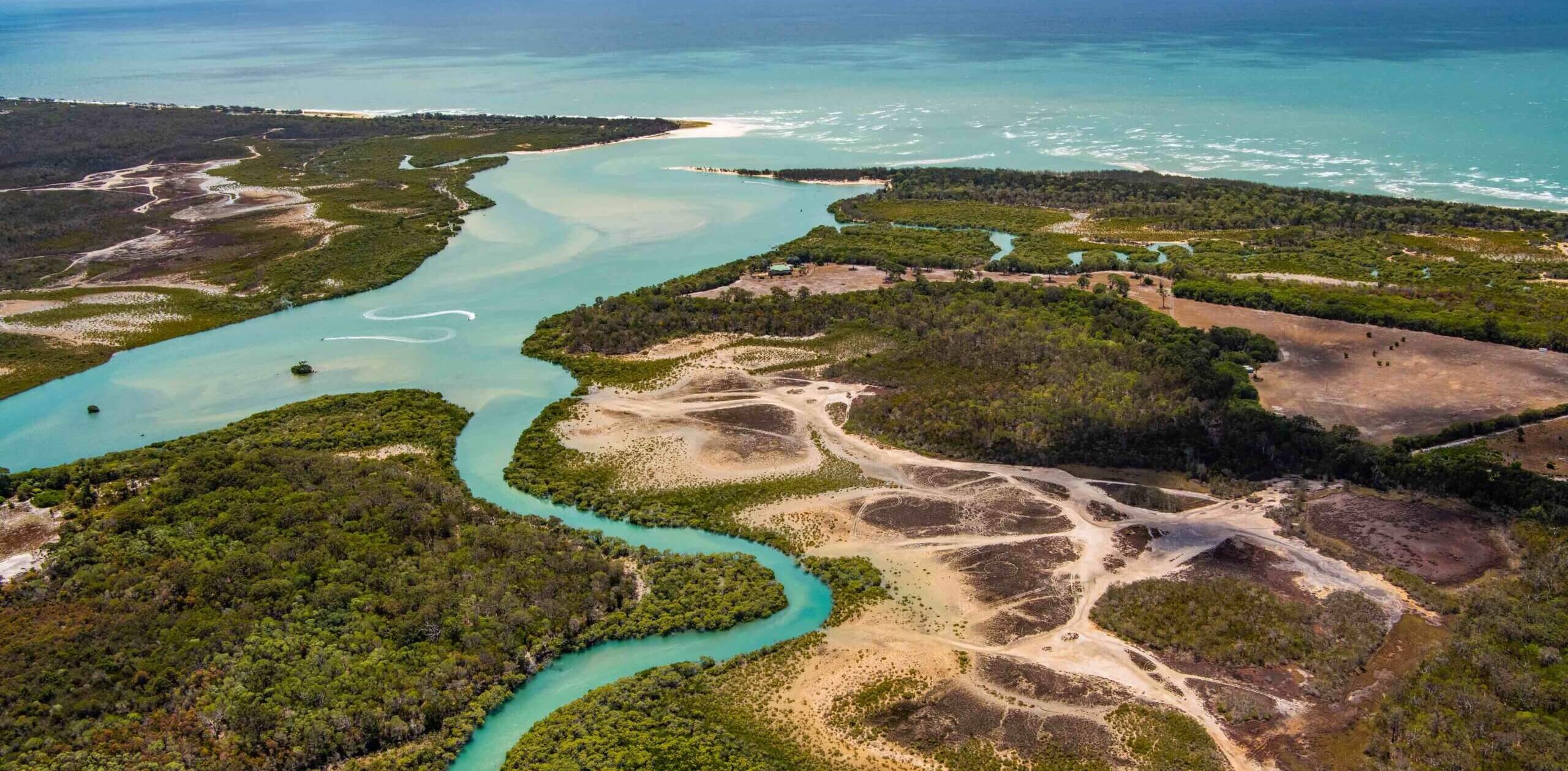 Aerial view of a winding turquoise river flowing through a lush landscape with patches of dense forest and open sandy areas. The river leads to a vast ocean, with the shoreline visible in the background.