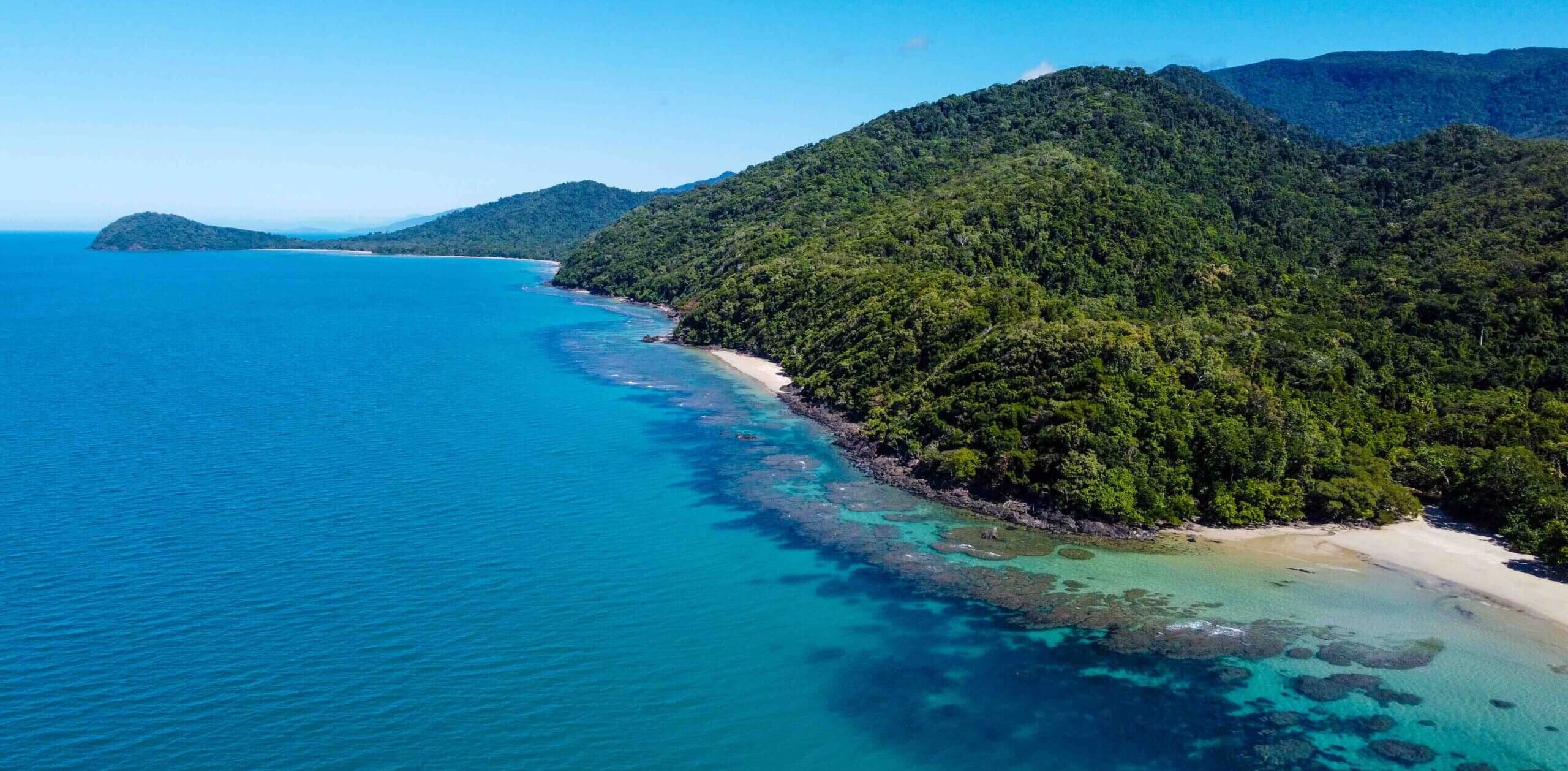 A vibrant aerial view of a coastline with lush green hills meeting clear turquoise waters. The shoreline is dotted with patches of coral and a small sandy beach, under a bright blue sky.