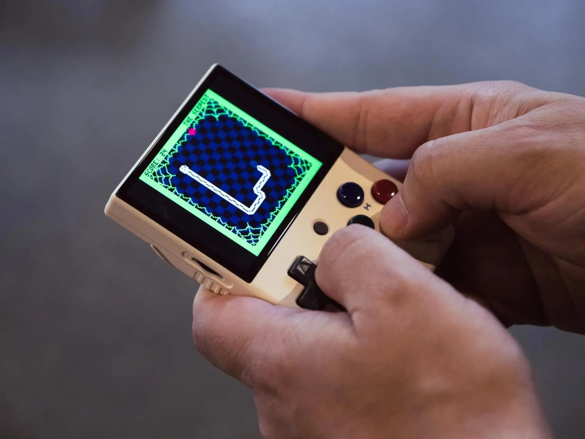 A person holds a small handheld gaming device, playing a retro-style snake game displayed on its screen. The person's thumbs are on the controls, and the background is out of focus.