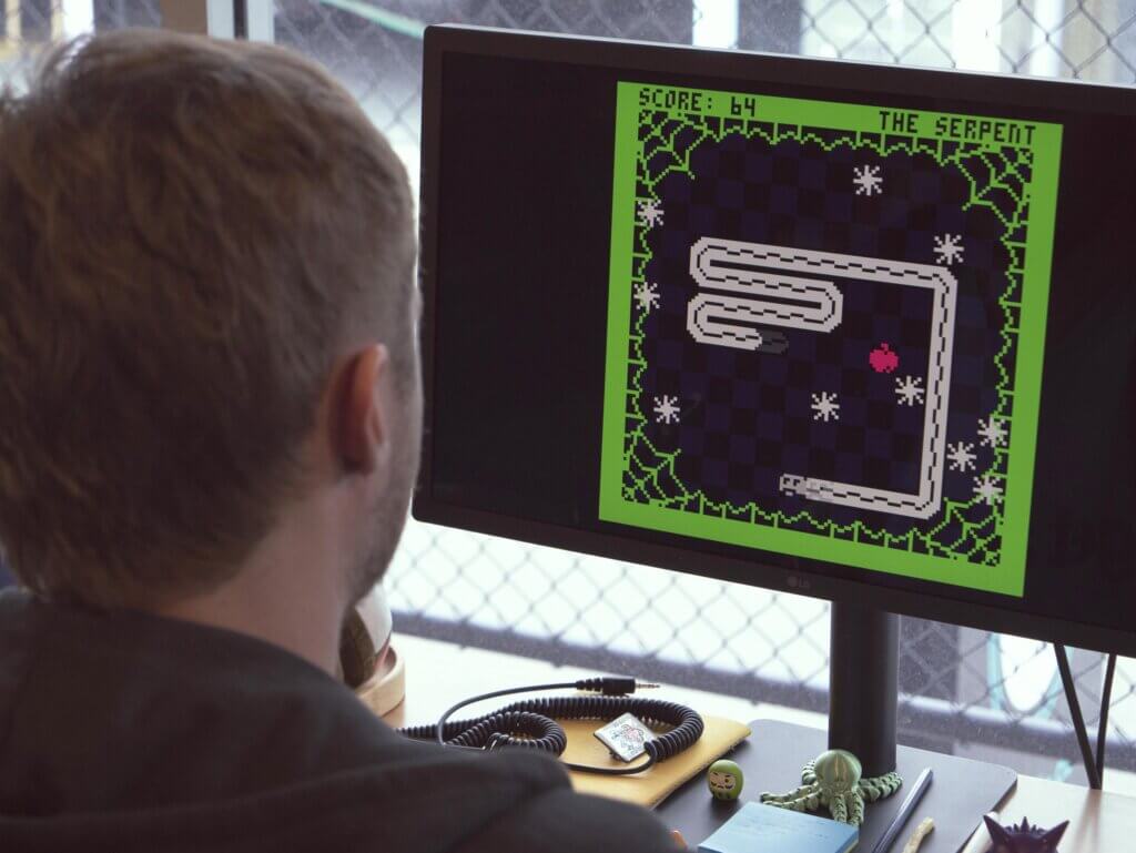 A person sits at a desk, facing a computer monitor displaying a retro-style snake game called "The Serpent." The game screen shows a winding snake and a score of 64.