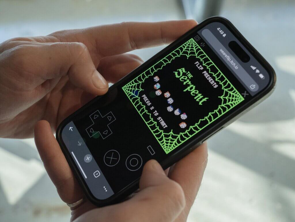 A person holds a smartphone displaying a retro-style game called "The Serpent," with a green spider web border and pixelated icons on the screen. On-screen controls for gameplay are also visible.
