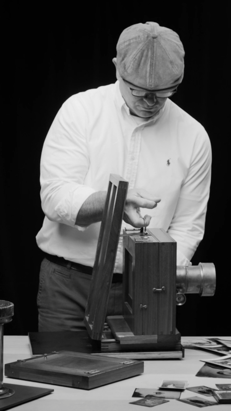 A person wearing a flat cap and glasses operates a vintage wooden camera in a black and white setting. Several photographs are scattered on the table beside them.