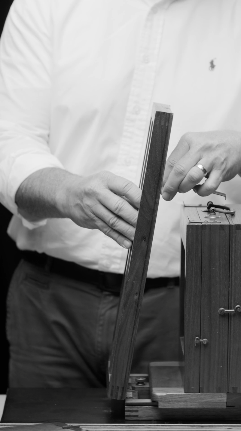 A black and white image of a man in a white shirt and dark pants interacting with a wooden object. His hands adjust a thin rectangular piece attached to another wooden structure, which has various small knobs and details.