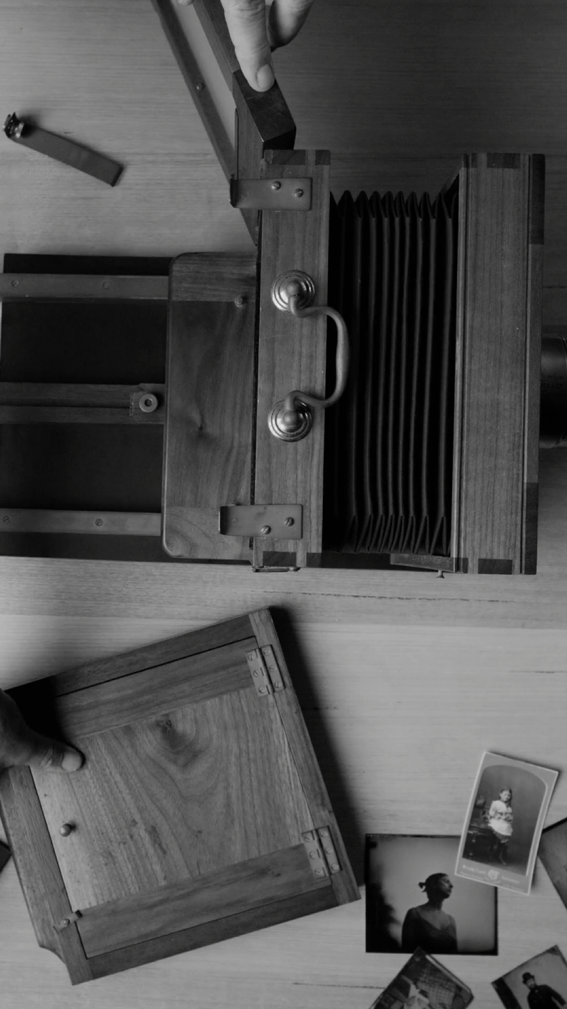 A black and white photo of a vintage camera with bellows, laid open on a wooden surface. Two hands are interacting with the camera. Around it, there are several small, old photographs scattered on the table.