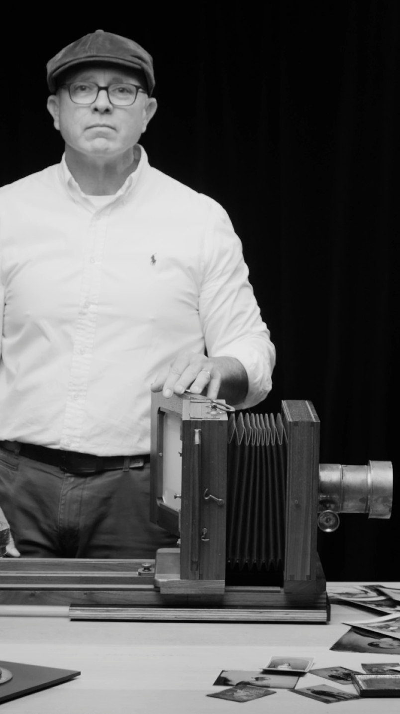 A man in a white shirt and cap stands next to a vintage camera with bellows on a table, surrounded by photographs. The background is dark, highlighting the historical camera.