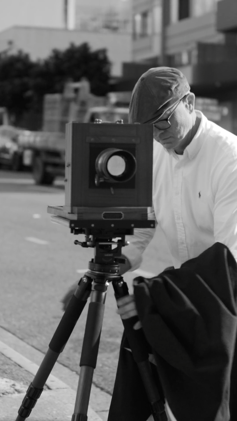 A person wearing a flat cap and glasses operates a large-format camera on a tripod, adjusting the camera while holding a dark cloth. They are outdoors, with blurred vehicles and buildings in the background. The image is in black and white.