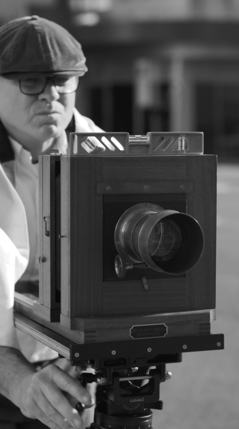 A person wearing a cap and glasses operates a large vintage camera with a wooden body and metal lens housing. The scene is in black and white, adding a classic feel.