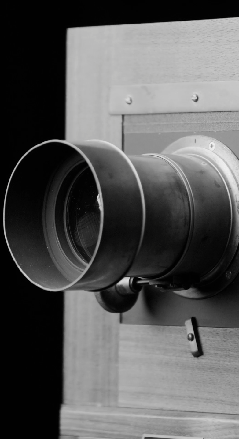 Close-up black and white image of an antique camera lens with a long, cylindrical shape mounted on a wooden box. The lens's detailed metal components and screws are visible against a dark background.