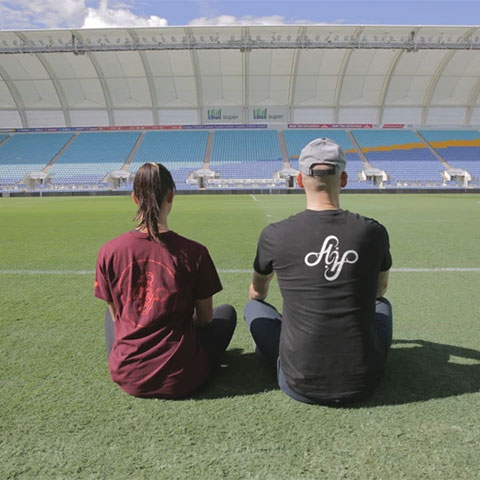 Two people sitting on grass facing a stadium