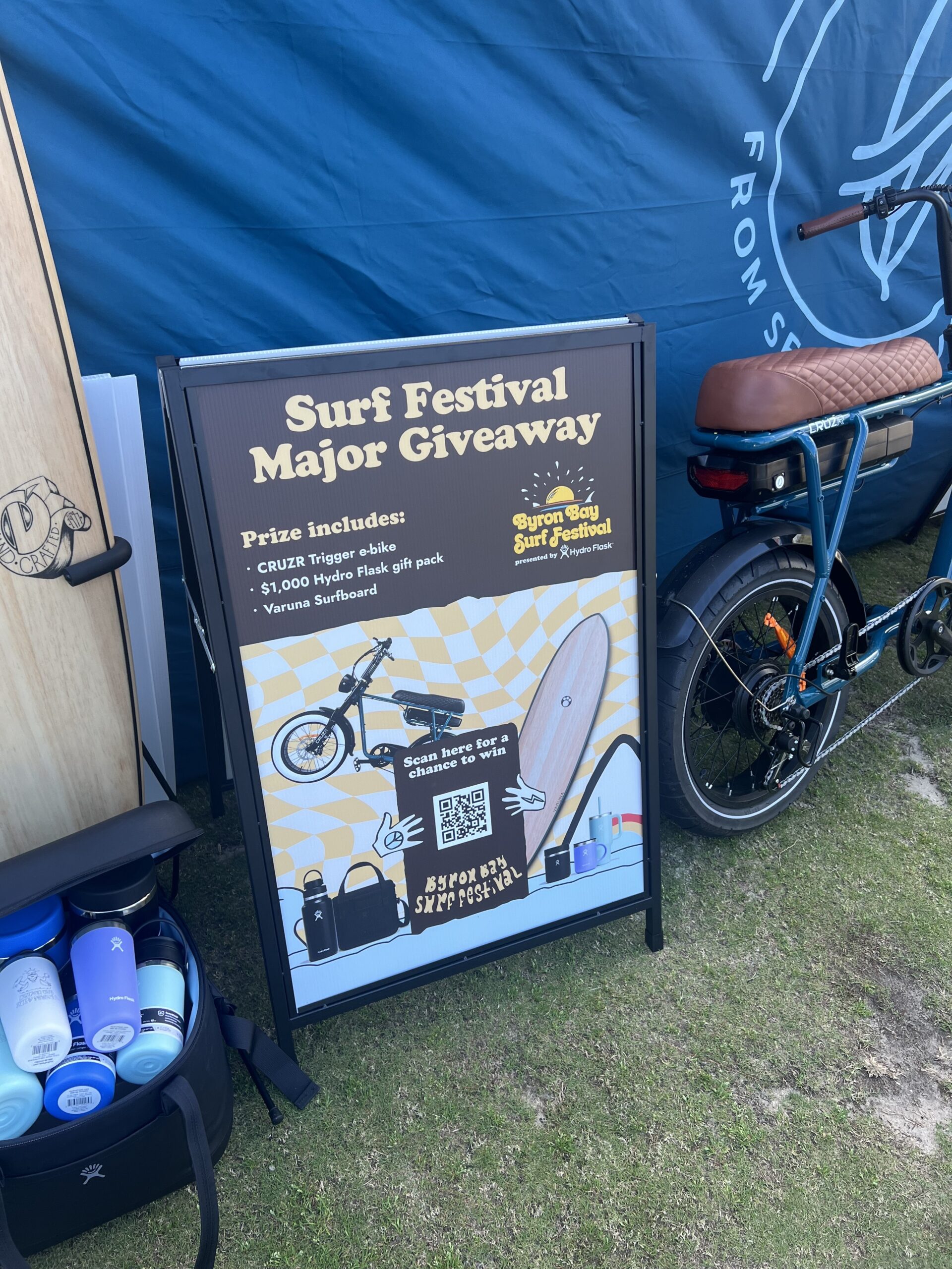 A standing sign at a surf festival advertises a major giveaway with prizes including an e-bike, Hydro Flask gift pack, and a surfboard. The sign features images of the prizes and is set next to a bike and surf gear.