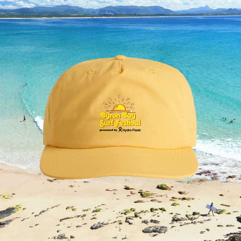 A yellow cap with "Byron Bay Surf Festival presented by Hydro Flask" written on it sits in front of a scenic beach with turquoise water, sandy shore, rocks, and distant surfers.