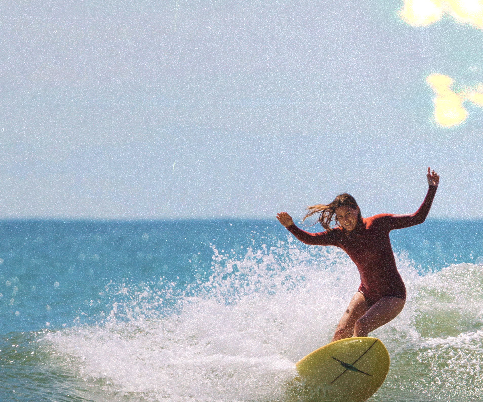 A person in a red wetsuit surfs a wave on a yellow surfboard, smiling and raising one hand. The ocean water splashes around them under a bright, sunny sky.