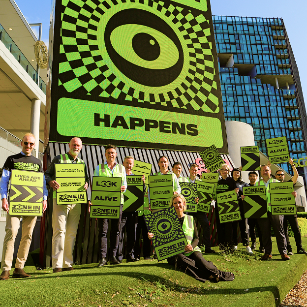 A group of people in safety vests hold neon green and black signs with eye graphics and road safety messages in front of a large digital display that reads "HAPPENS" and shows a large eye graphic.