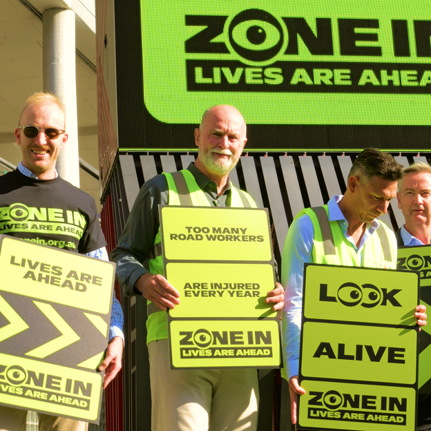 Four men stand outdoors holding bright yellow-green ZONE IN road safety signs with messages like "LIVES ARE AHEAD," "TOO MANY ROAD WORKERS ARE INJURED EVERY YEAR," and "LOOK ALIVE." A large sign is visible behind them.