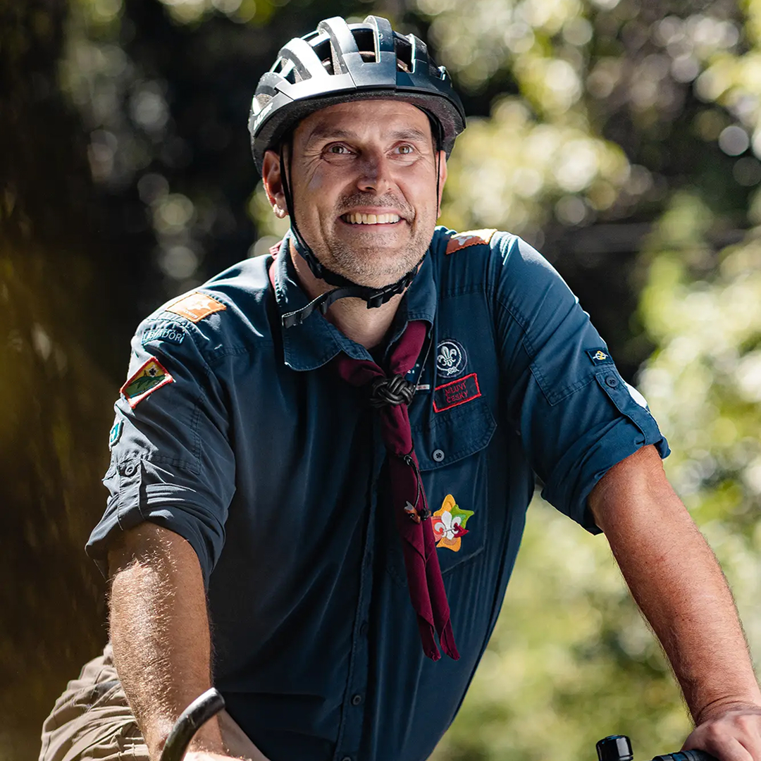 A man wearing a bike helmet and a Scout uniform smiles while riding a bicycle outdoors, surrounded by greenery and sunlight filtering through the trees.