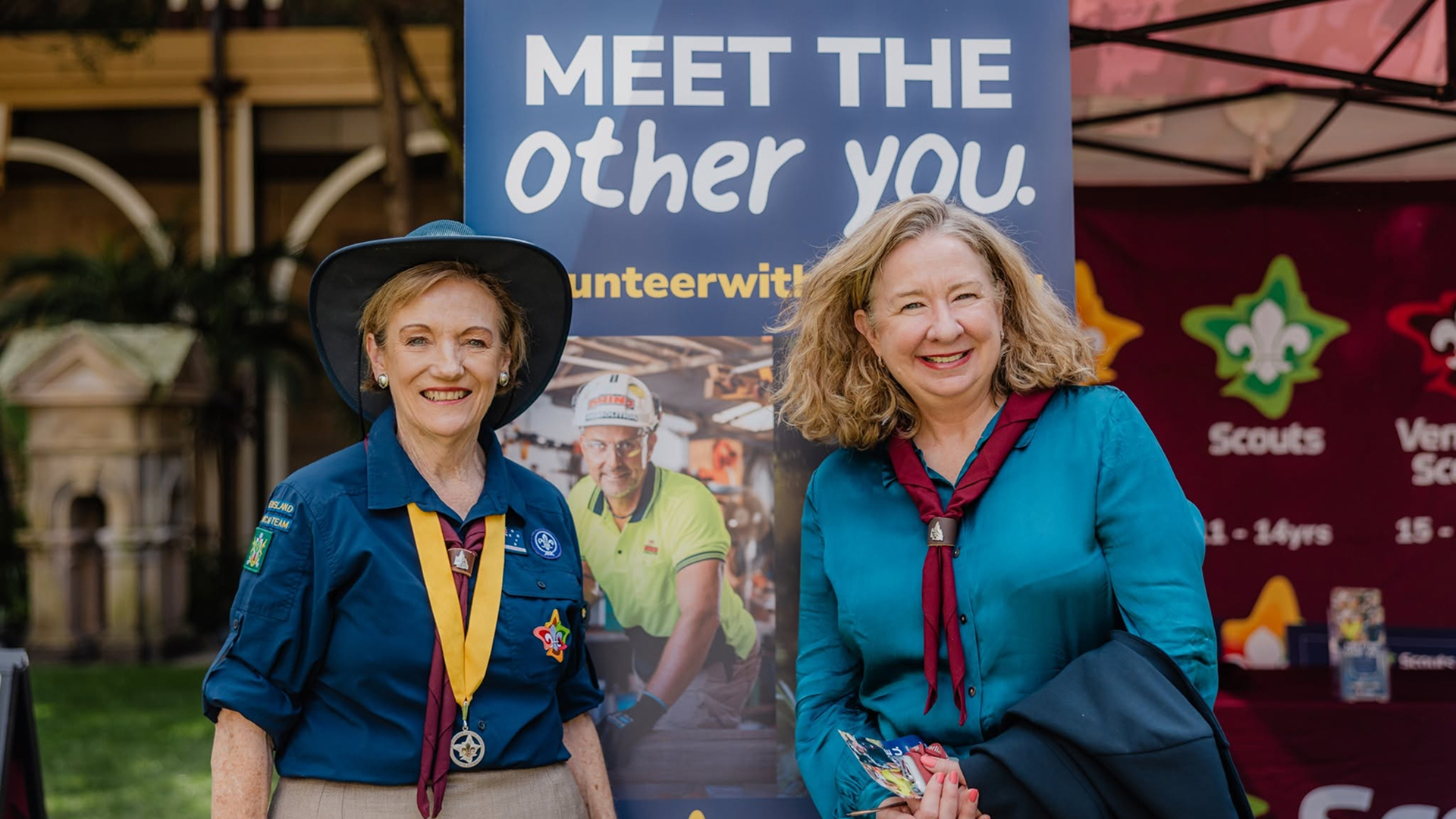 Two women smile in front of a sign that says "Meet the other you." One wears a scout uniform and hat with badges and a yellow scarf; the other wears a teal blouse with a maroon scarf. Scout banners are visible in the background.