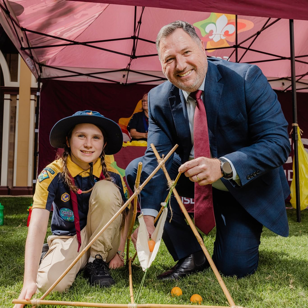 A smiling adult in a suit kneels next to a child in a scout uniform. They are outdoors on grass, under a maroon canopy, working together on a wooden structure with small orange balls.