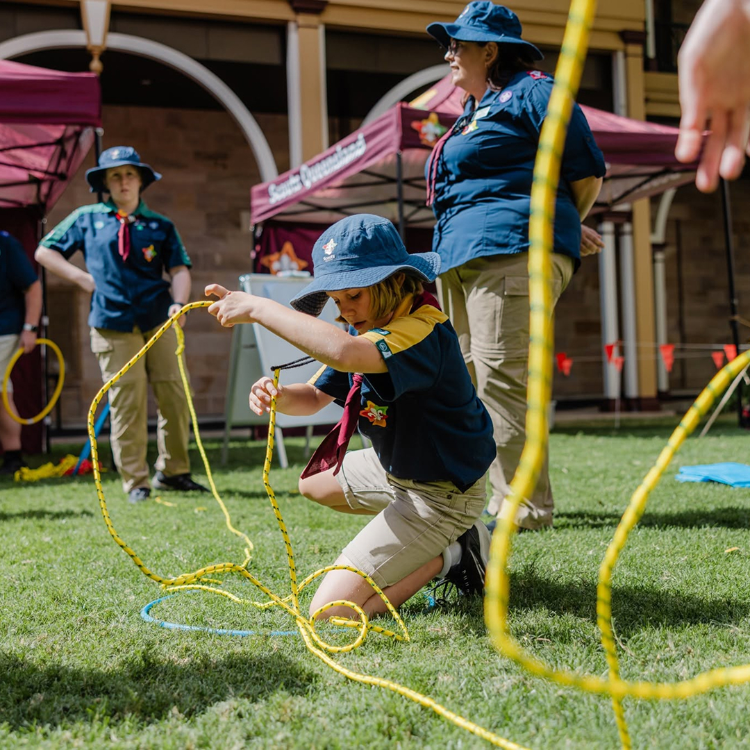 A child in scout uniform kneels on grass, handling yellow rope, with other scouts and an adult leader nearby. They are outdoors near tents and a building, participating in an activity on a sunny day.