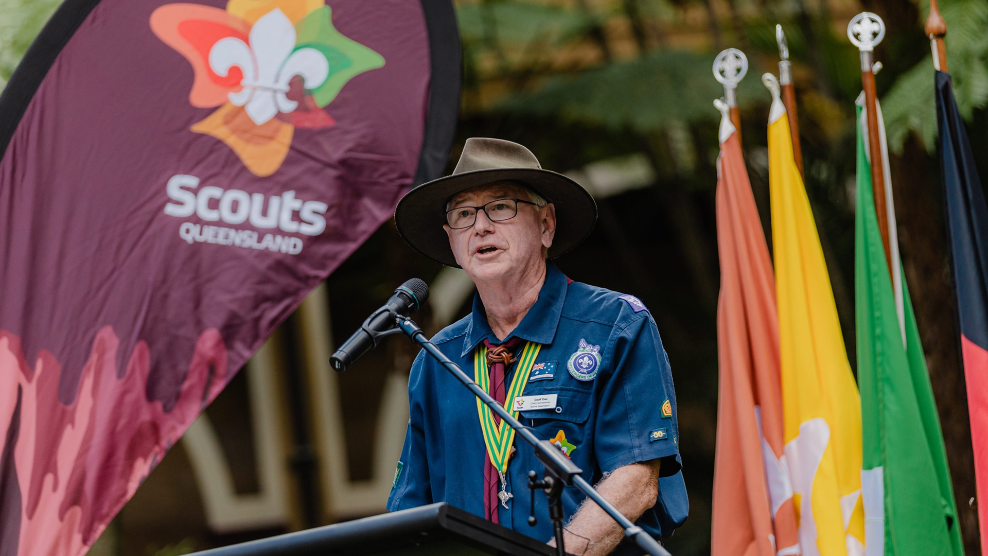 A man in a scout uniform and hat speaks at a podium with a microphone, beside a "Scouts Queensland" flag and several colorful flags in the background.