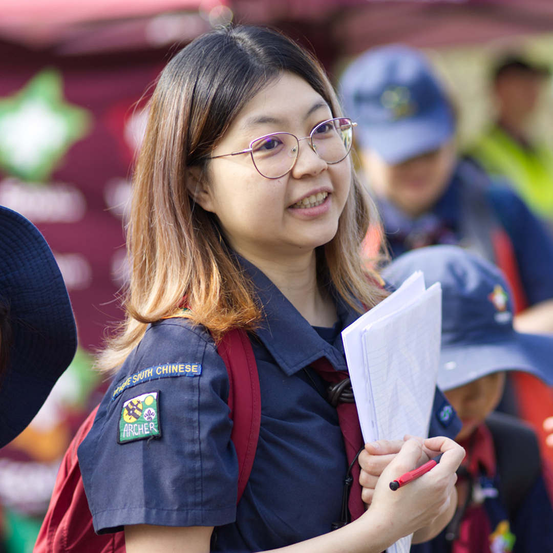 A young person with glasses and shoulder-length hair wears a navy scout uniform and red backpack, holding papers and a pen outdoors. Others in similar uniforms are visible in the background.