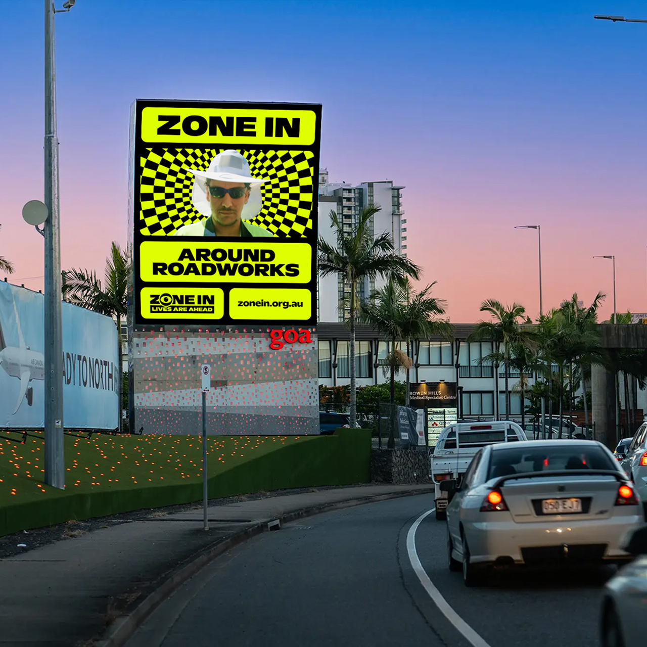 Digital billboard beside a busy road at sunset reads “ZONE IN AROUND ROADWORKS” with a photo of a man in a hard hat and sunglasses. Cars drive past palm trees and buildings under a pink and blue sky.