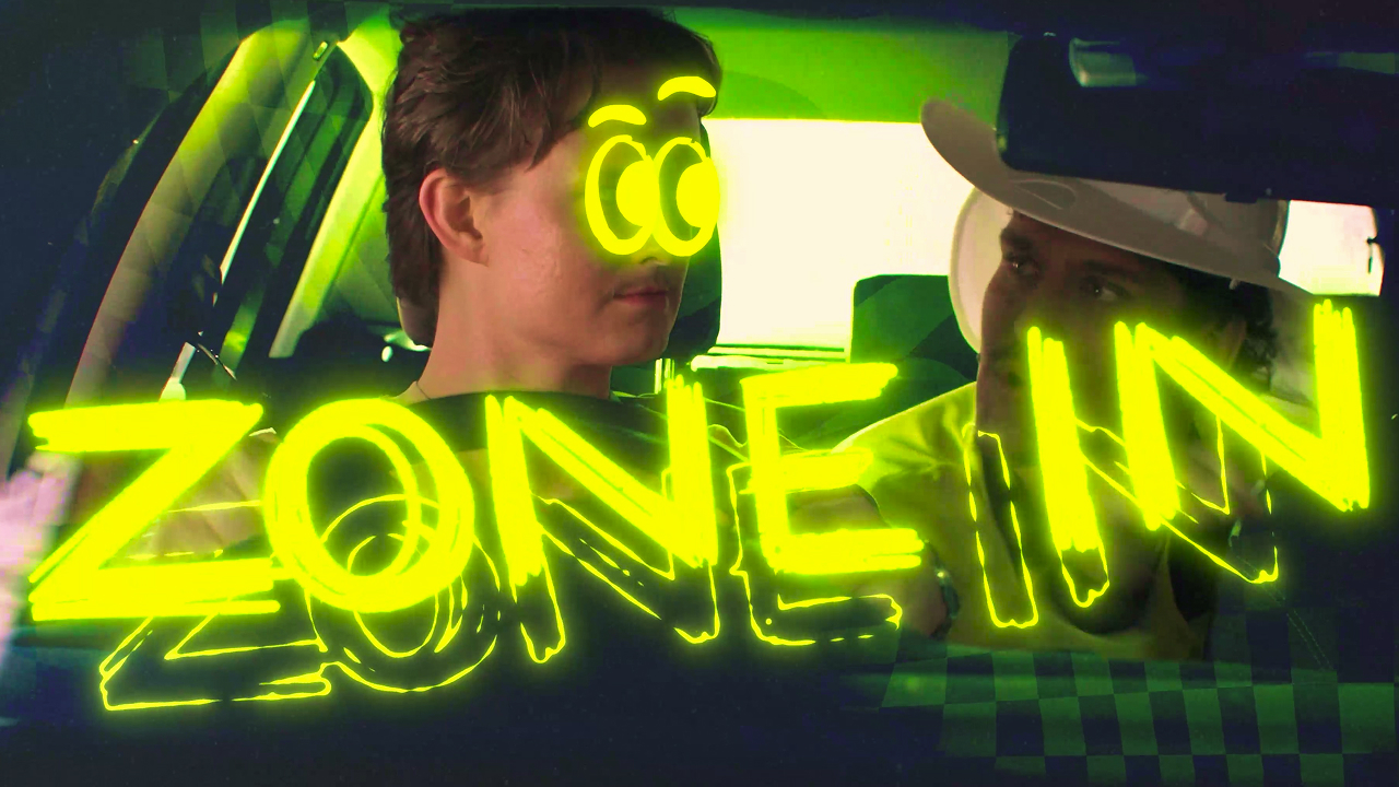 Two people sit in a car, one with drawn neon yellow cartoon eyes and eyebrows. Bright neon yellow text reading "ZONE IN" is overlaid across the image, giving it a surreal, stylized look.